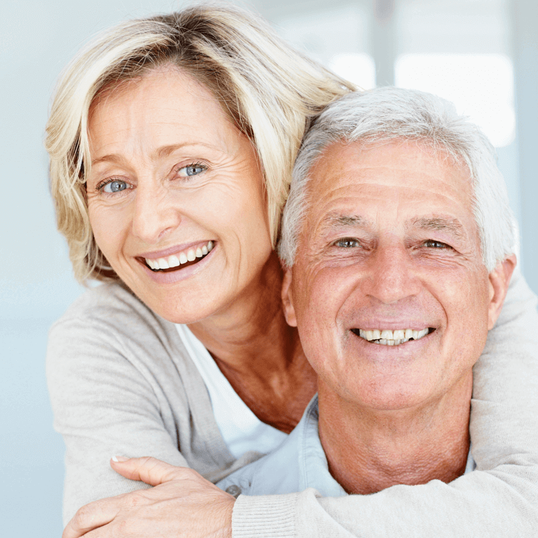 Elderly women holding elderly man from behind while smiling with dental implants